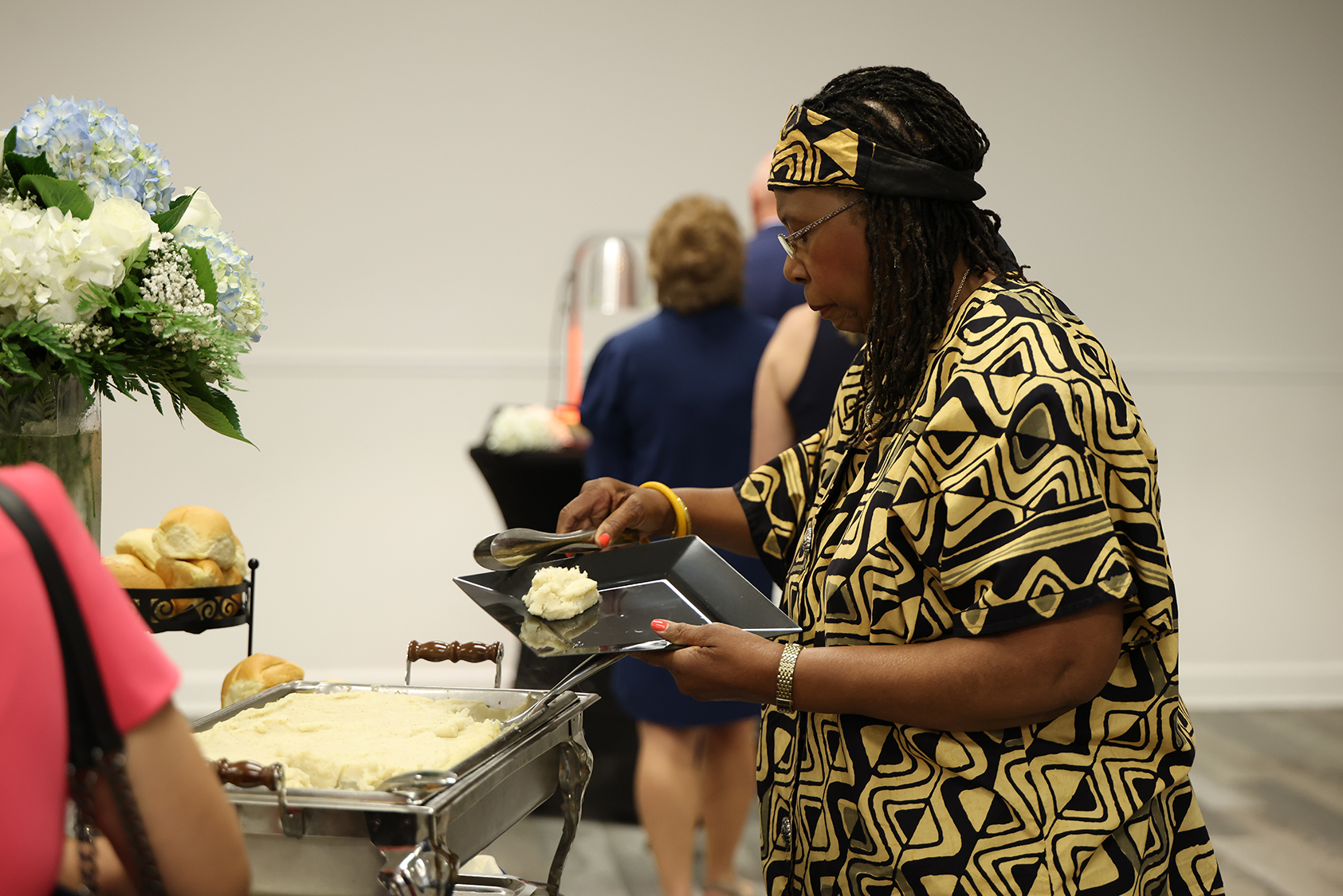 Person at a table with a food tray. Person in black and gold patterned top serving food.
