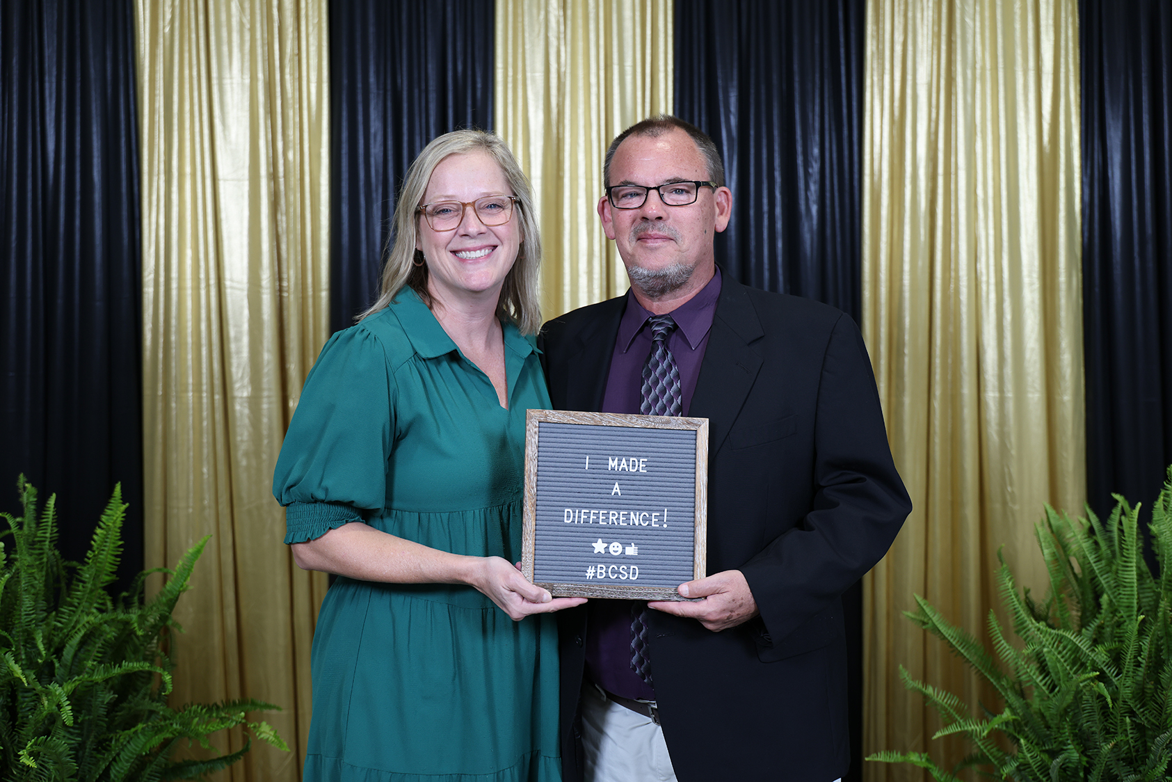 A woman and man in green and black suits hold a plaque in front of a golden backdrop.