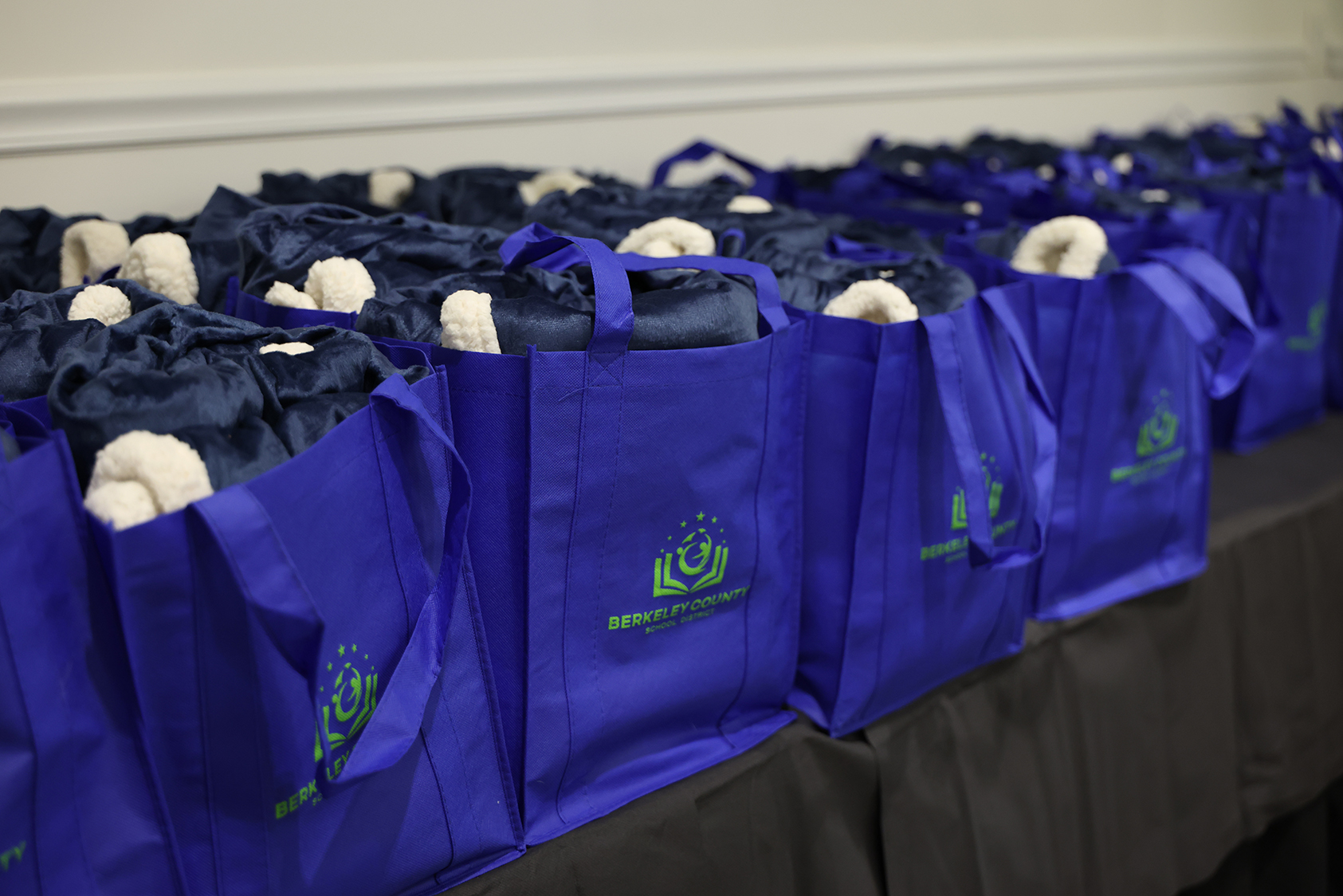 Row of blue bags with white cloths and green logos on a table against a white wall.