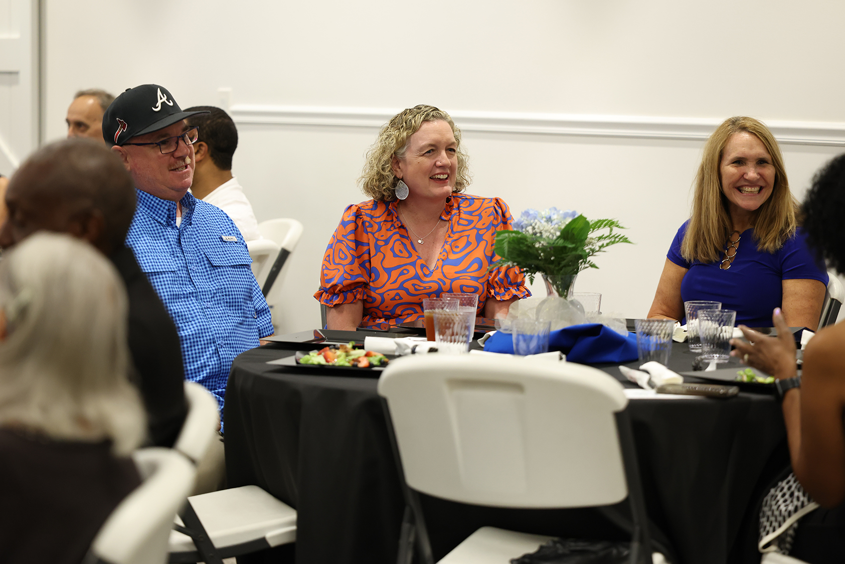 A group of people sit around a table, smiling and talking. One woman wears an orange floral top.