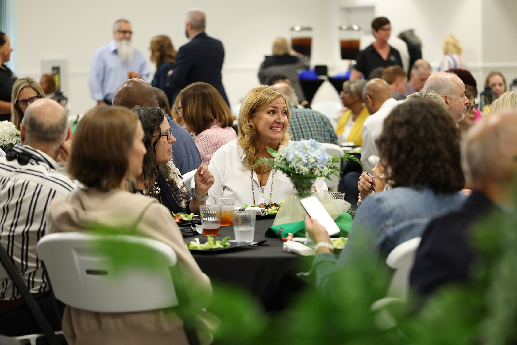 A group of adults, some seated at tables, enjoy a meal at an event. One woman smiles at the camera.