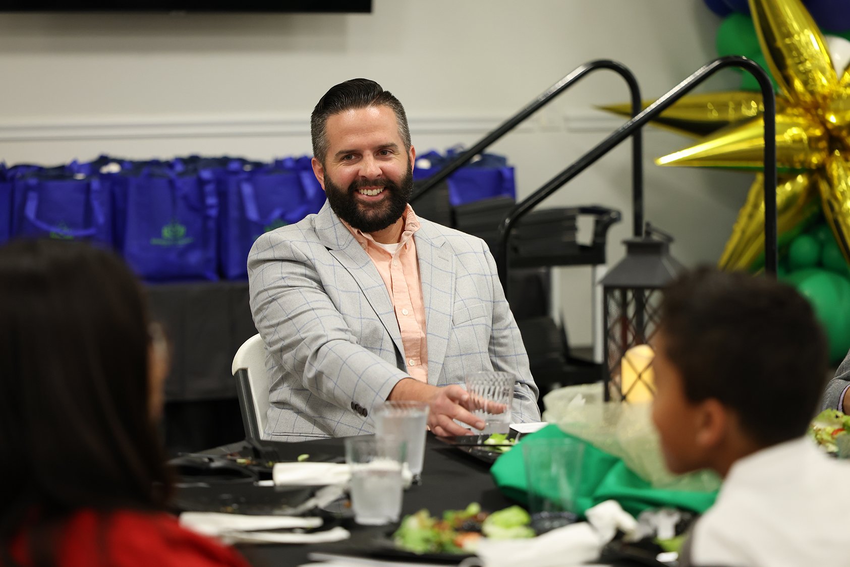 A man in a gray suit and beard sits at a table with others, gesturing while talking.