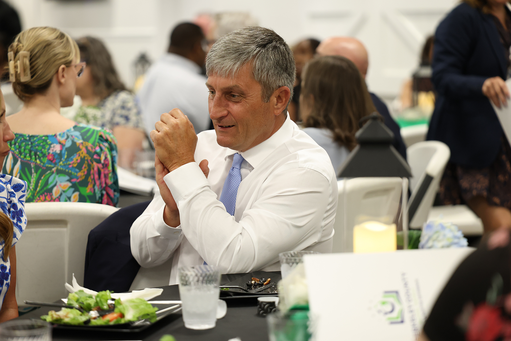 A man wearing a white shirt and blue tie sits at a table, smiling, with a plate of food.