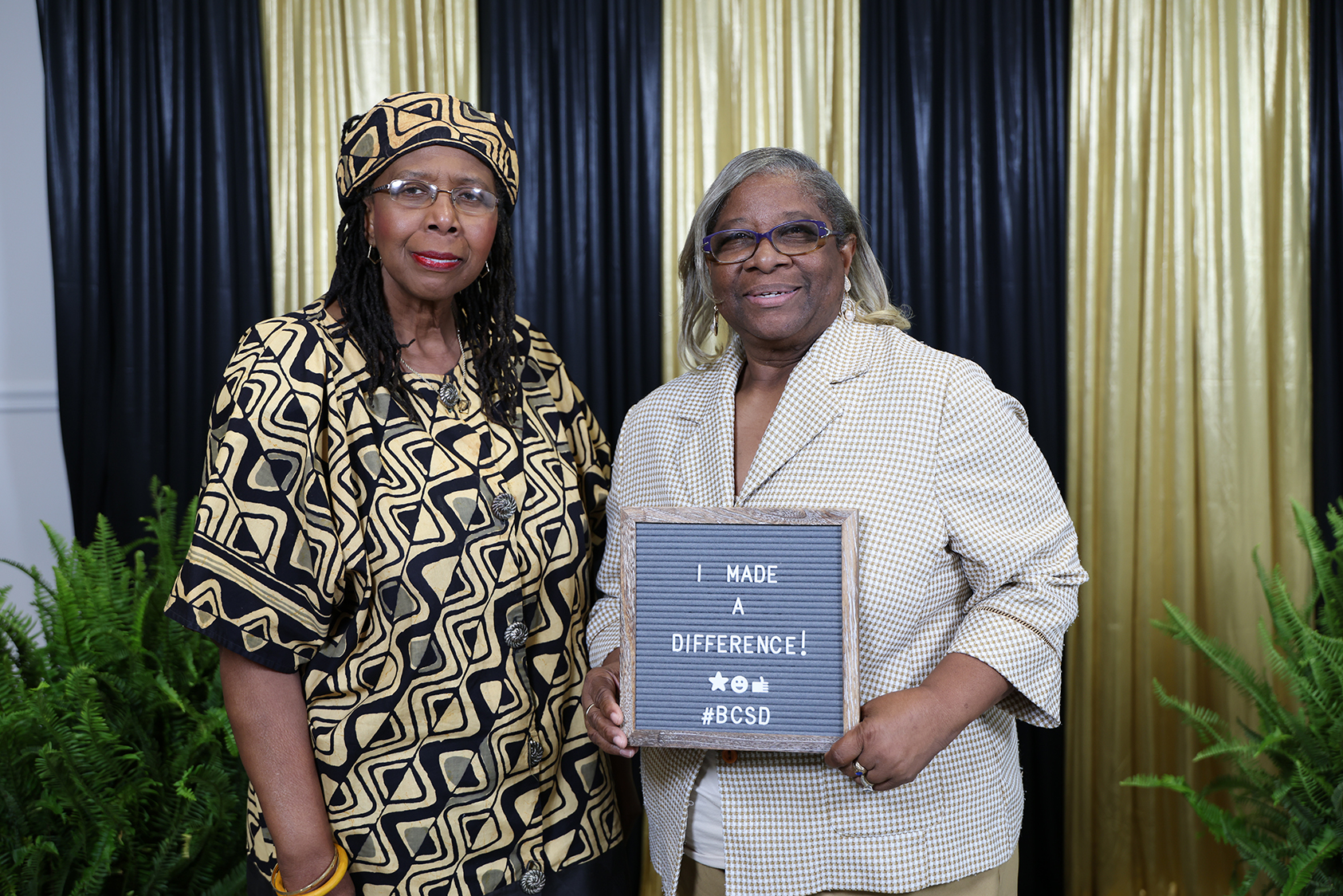 Two women pose for a photo in front of a black and gold curtain backdrop, one holding a plaque.