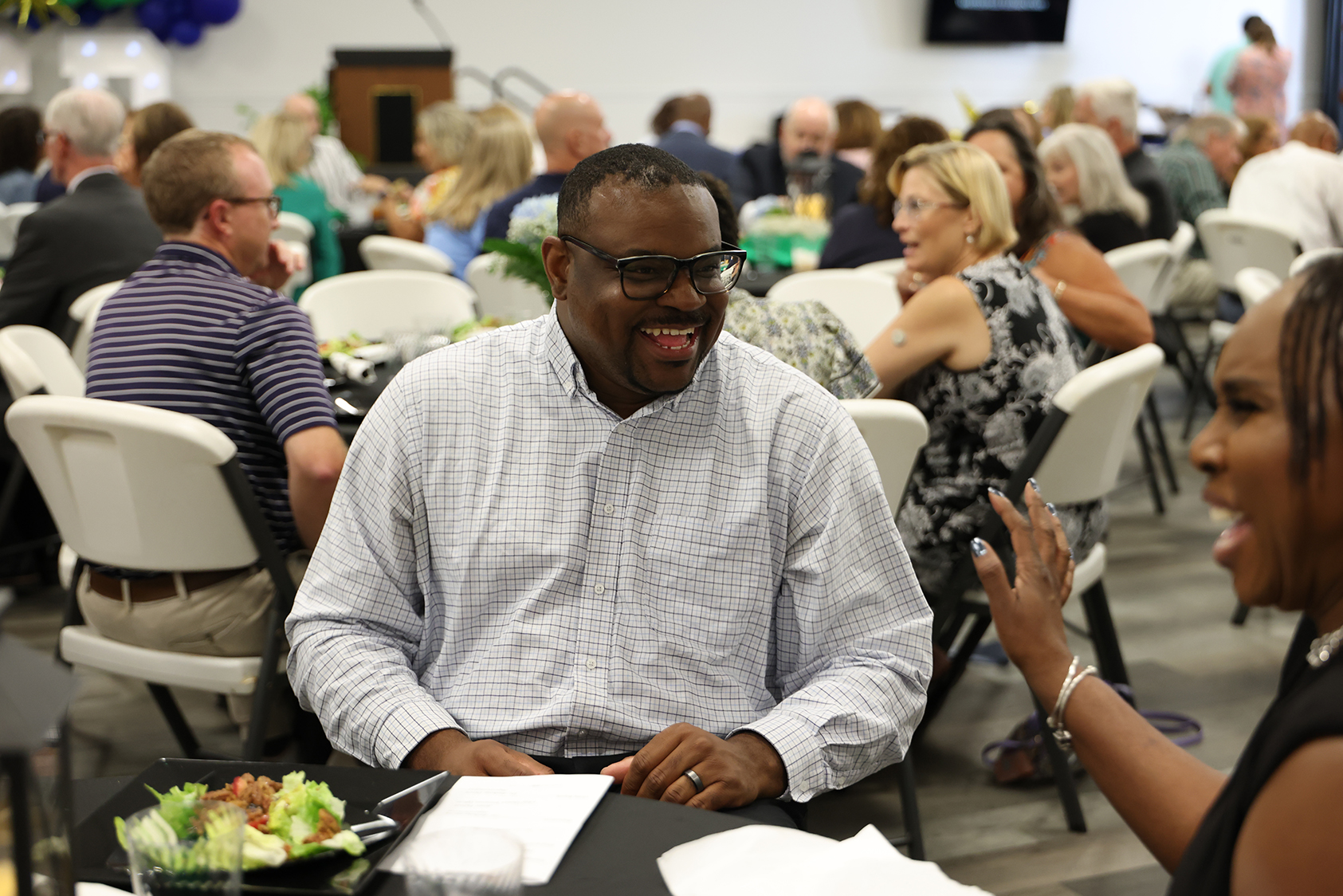 A man in glasses and a white shirt smiles at a woman seated at a dining table.