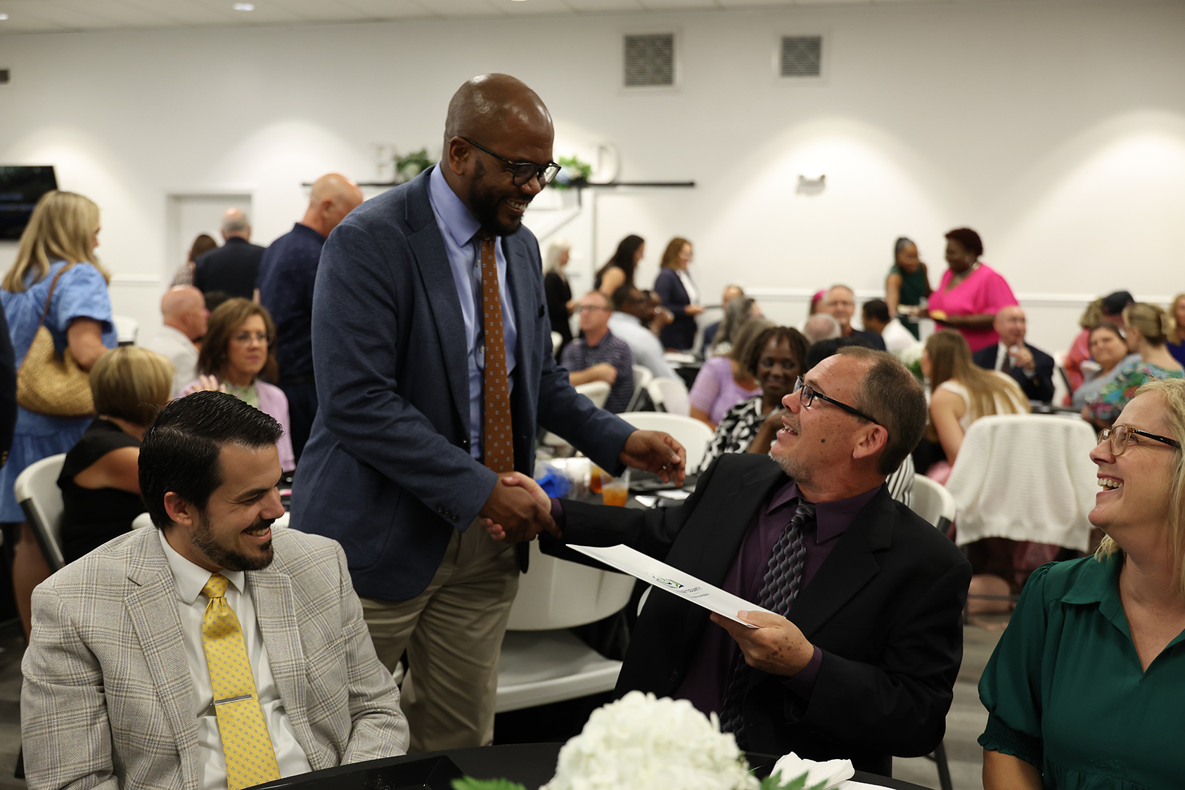 An indoor event with people in business attire. A man hands a paper to another man seated at a table with flowers.