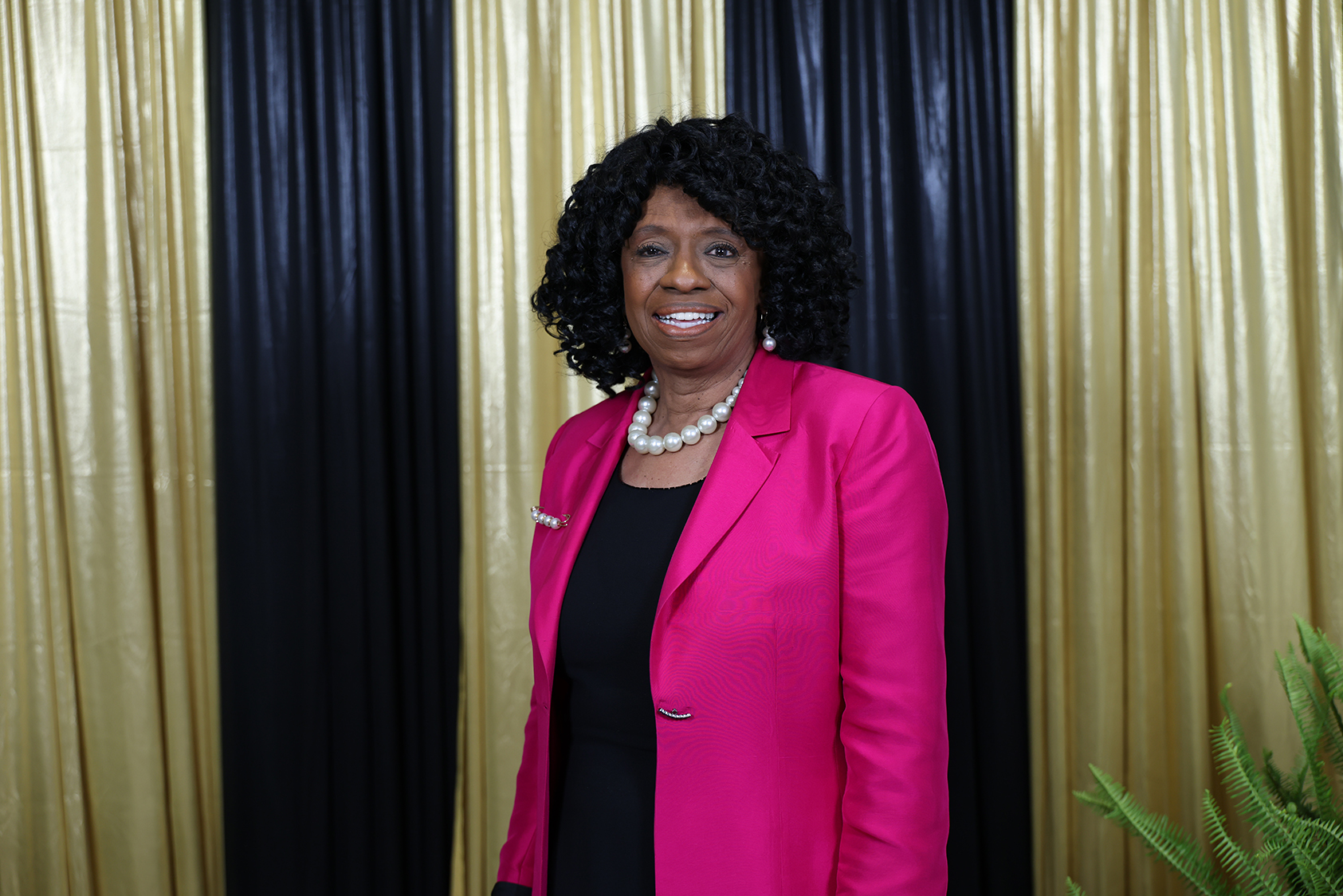 A woman with curly hair wearing a pink blazer, black top, and pearl necklace stands before a gold and black curtain backdrop.