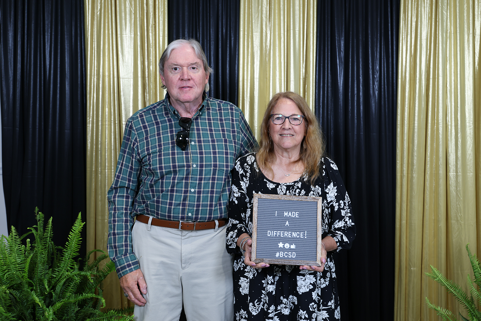 A man and woman stand before a gold and black curtain backdrop. The woman holds a framed plaque.