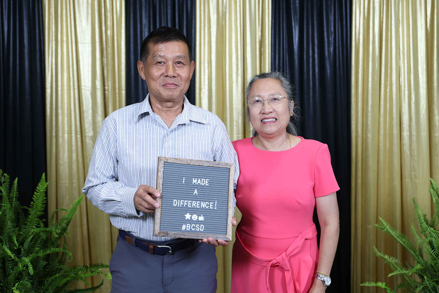 Man and woman standing together, smiling, with a blackboard between them. Behind them, a black and gold curtain.