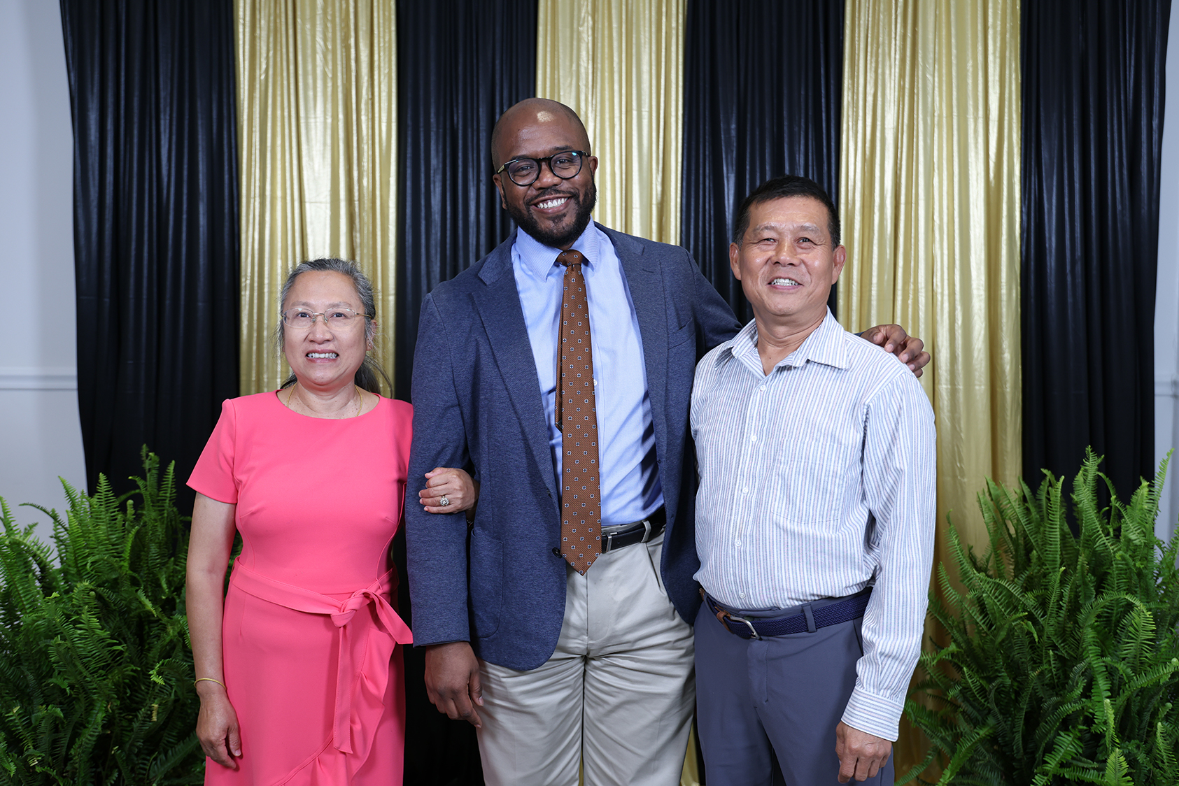 Three people standing in front of a decorative backdrop. A woman in pink, a man in gray, and a man in a suit.