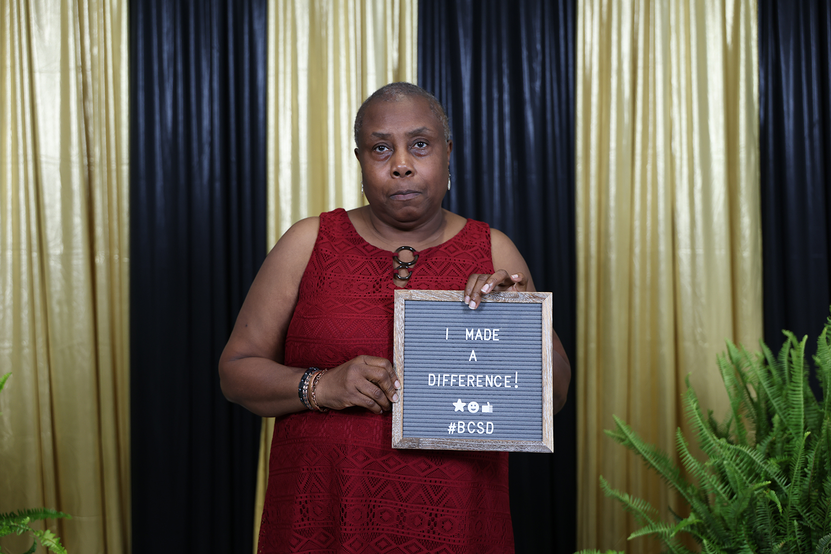 A woman in a red dress holds a small chalkboard saying "I MADE A DIFFERENCE" with a plant behind her.
