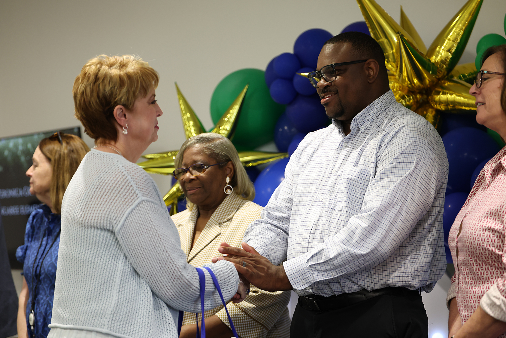 A man and a woman shake hands while two other women stand nearby. Behind them, colorful balloons are present.
