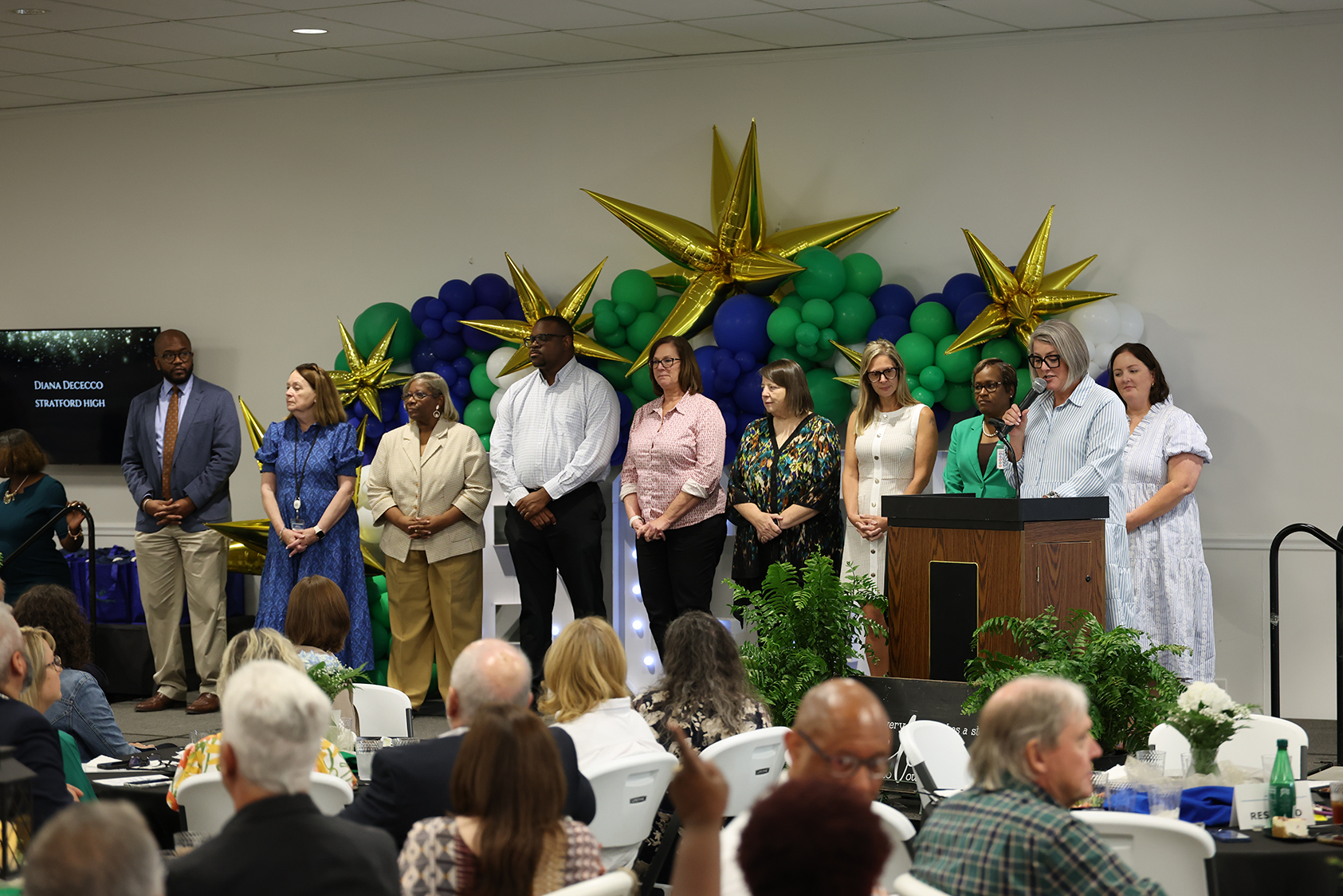 A group of people stands on stage with large star balloons in the background. An audience sits in chairs.