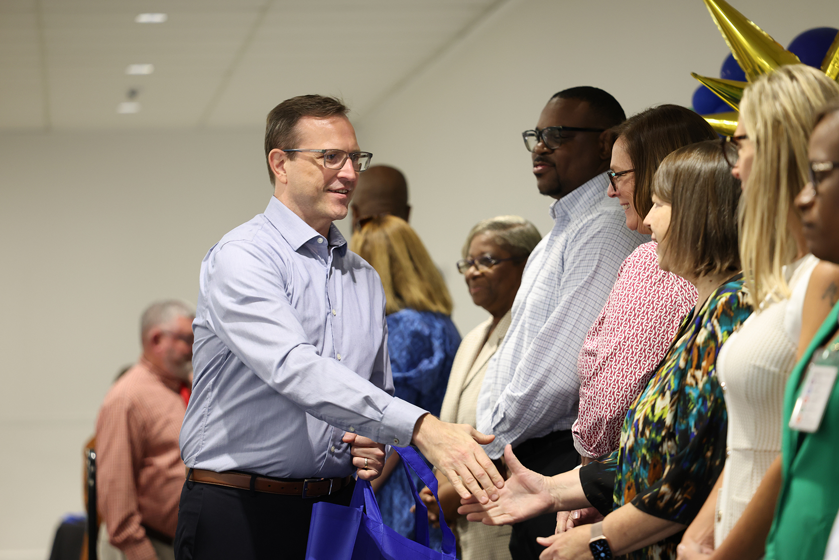 A group of people, including a man in a blue shirt, stand in a room. Some hold hands.