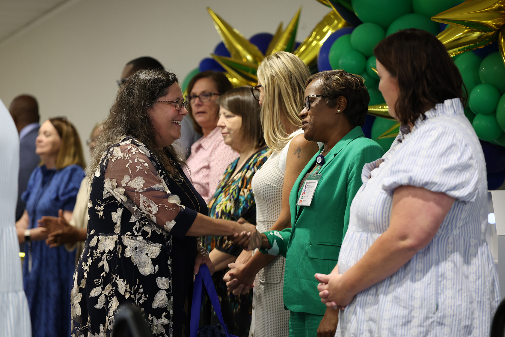 A group of people shaking hands in a room, with some wearing glasses and some holding balloons.