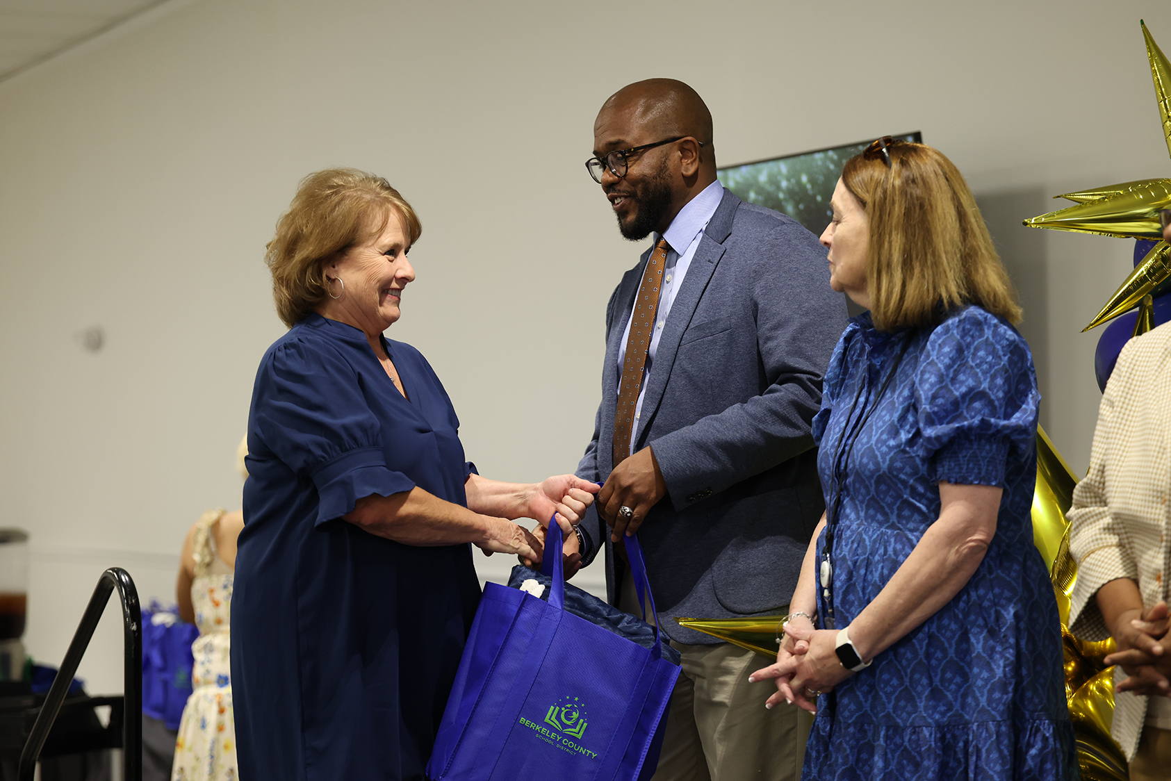 Three adults in blue dresses and a gray suit stand together. Two women hold hands, smiling. The man hands something to the woman on the left.