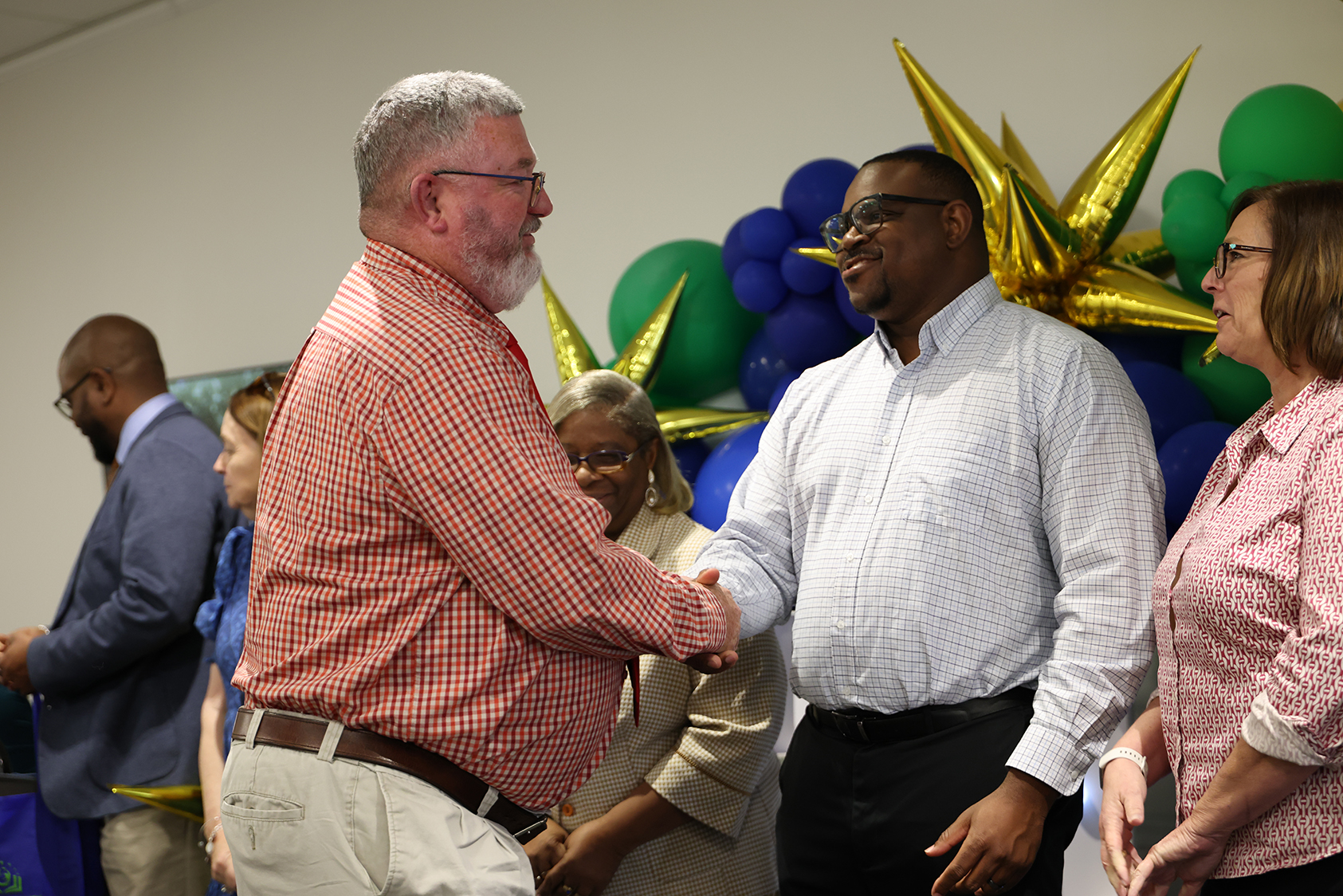 A group of people are standing, with two men shaking hands. One man wears a checkered shirt, and the other a white shirt.
