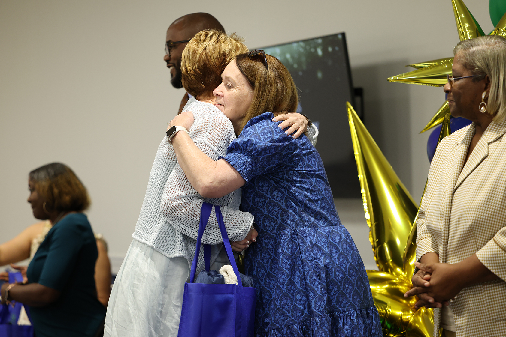 A group of people, some hugging and others standing, in an indoor space with a wall backdrop and decorative balloons.