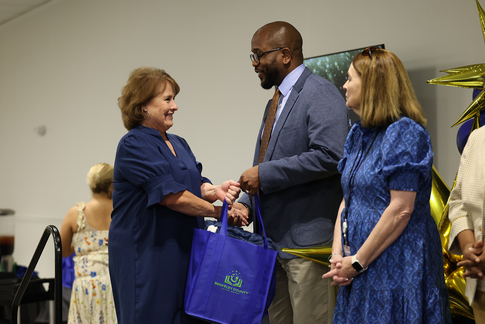 Three people in a room exchange a handshake. A woman holds a blue bag. Behind them are balloons and a table.