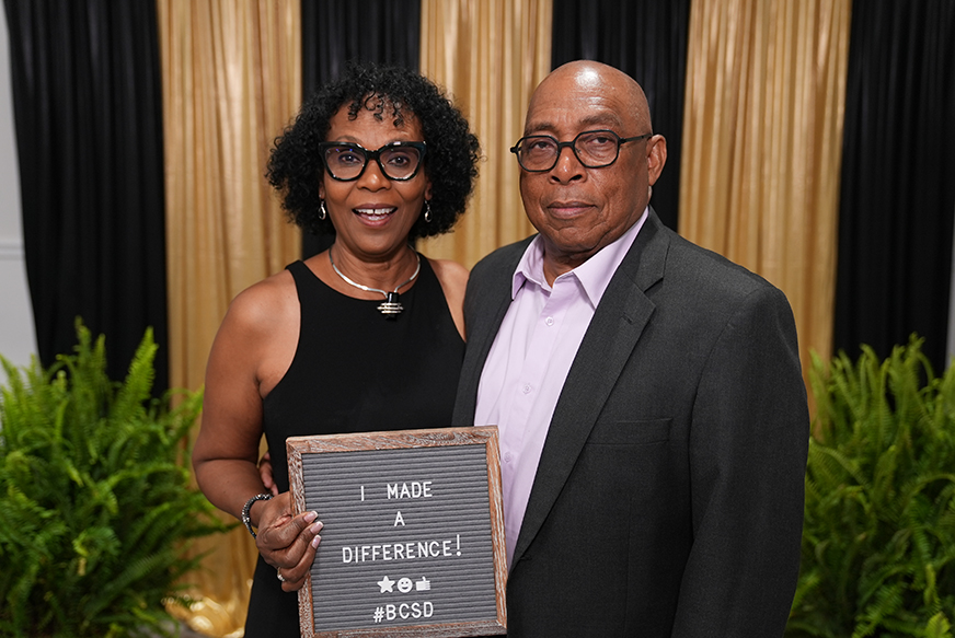 Man and woman holding a plaque with "I Made a Difference!" and a social media hashtag, standing with plants.