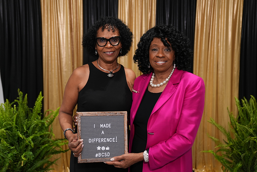 Two women stand next to each other, one holding a chalkboard sign saying, "I MADE A DIFFERENCE!" against a backdrop of plants and a black and gold curtain.