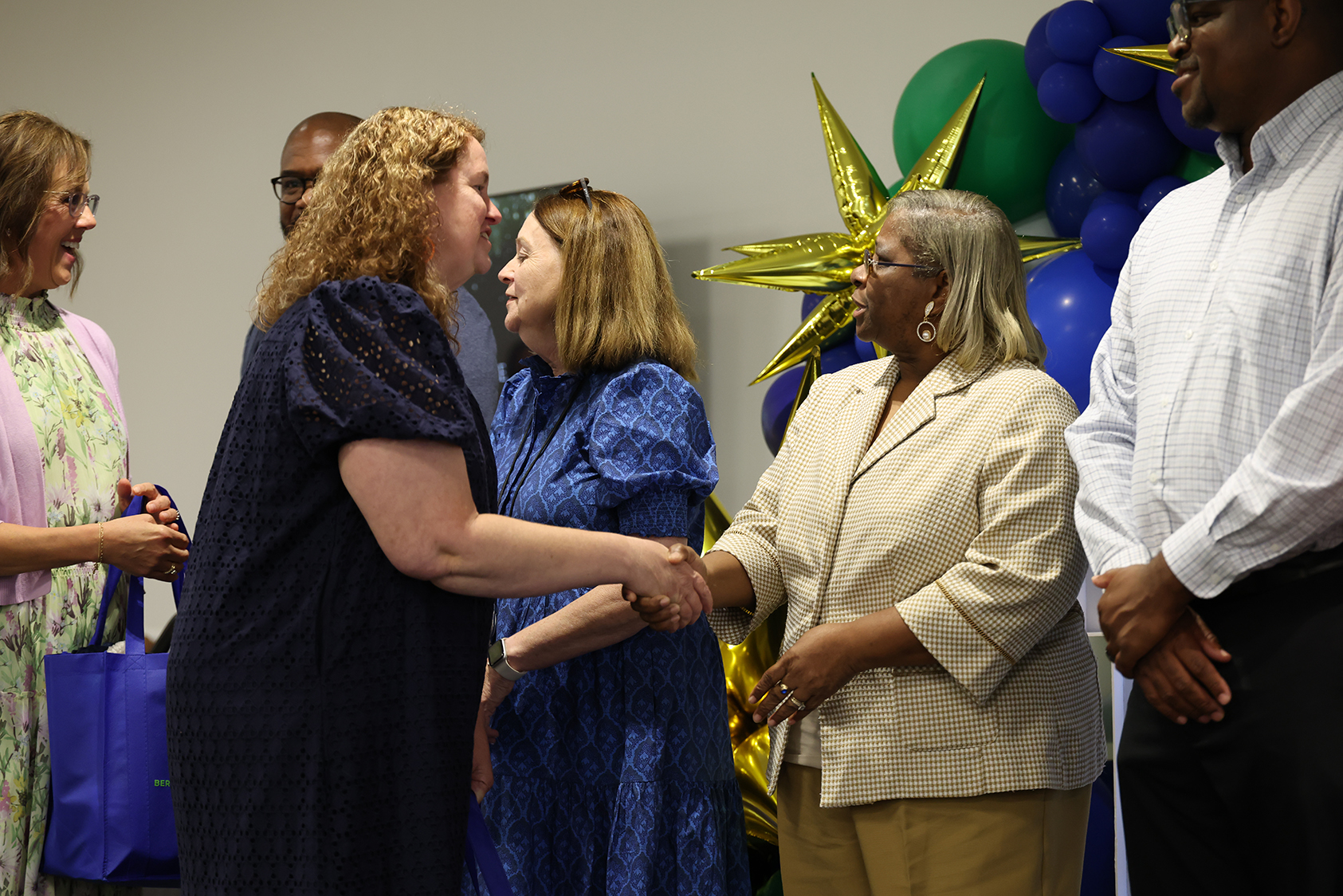 Five people stand close to one another. Two women shake hands. Two women wear blouses and skirts. One woman wears a blazer. Balloons behind them.