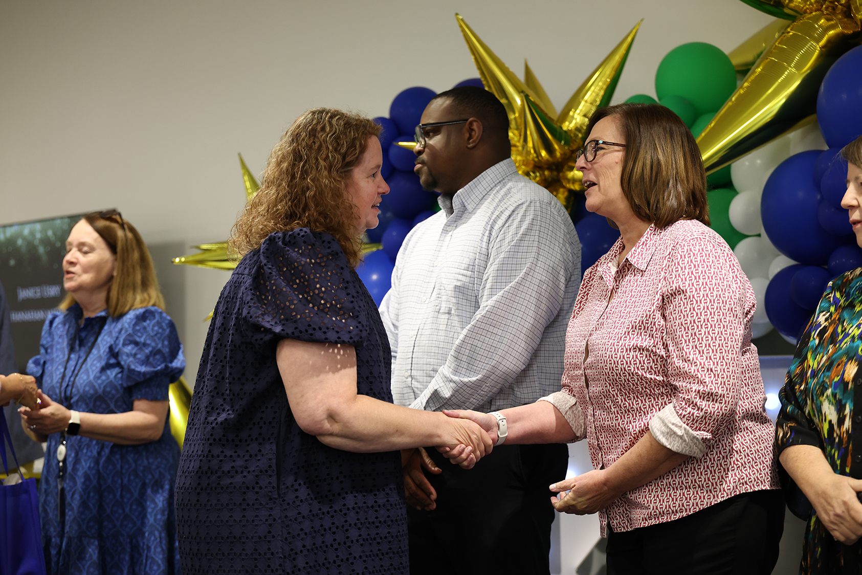 People stand with hands on shoulders, shaking hands in a room with a wall of balloons.
