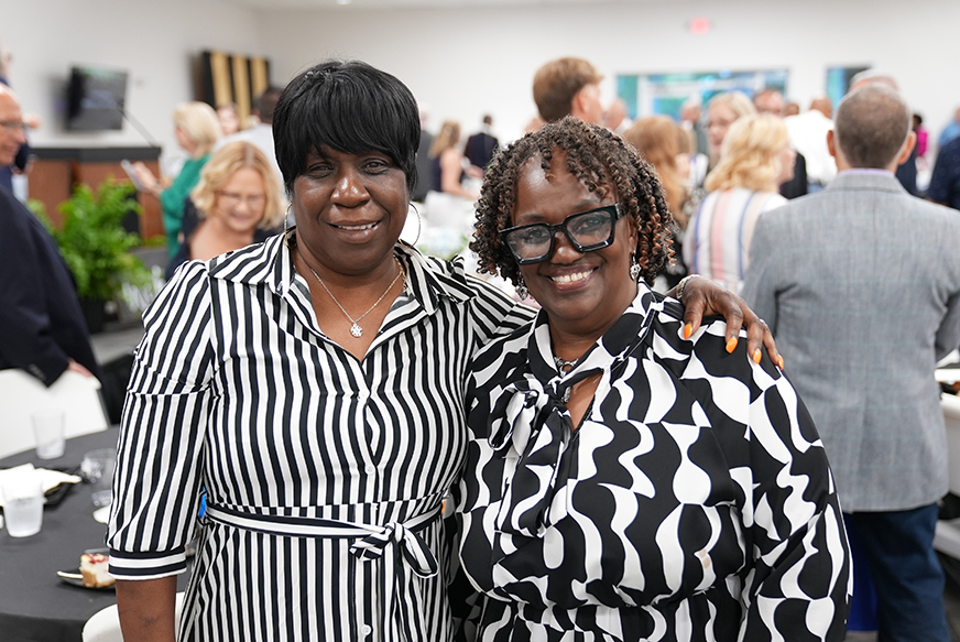 Two women, one with glasses and a patterned top, and the other with a striped top, pose together with arms around each other.