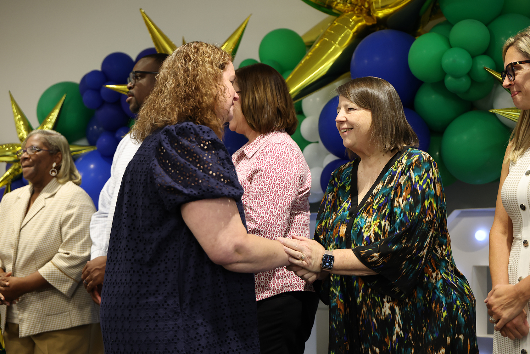 Group of people in business attire standing in front of balloons, two women in the front shaking hands.
