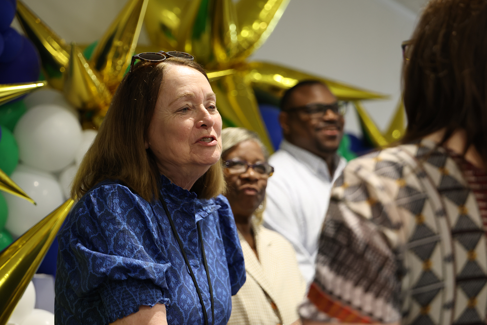 A woman in blue speaks into a microphone, with two others behind her, near a backdrop of balloons.