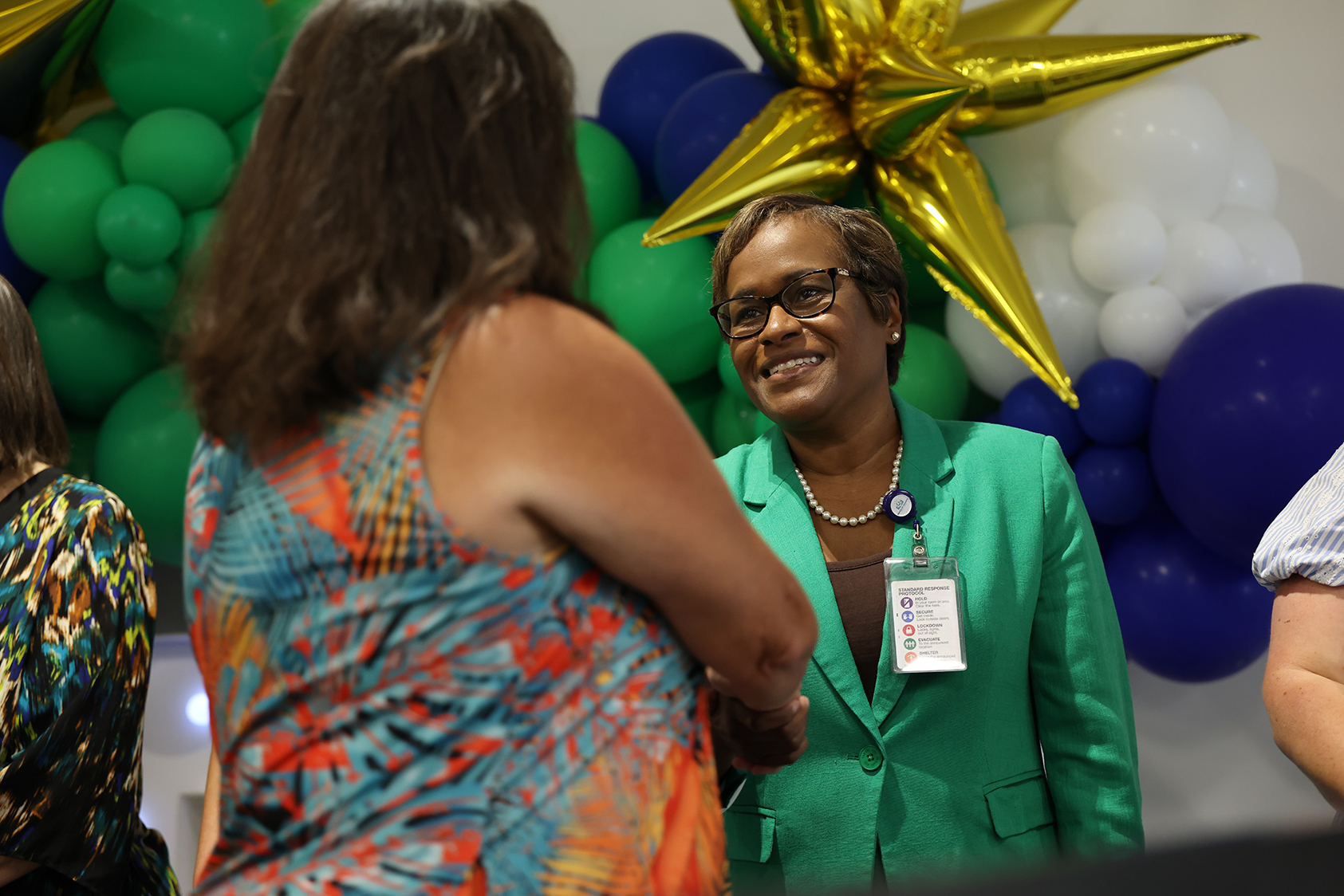 Two women converse near colorful balloons and a gold star. One wears a green blazer, the other a colorful dress.