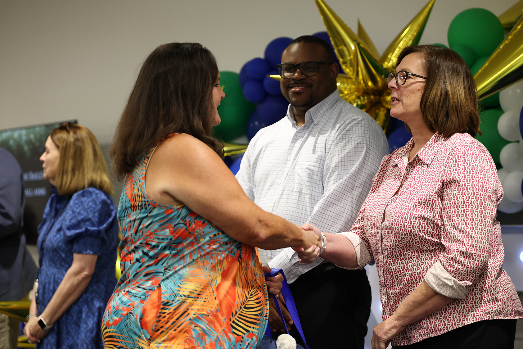 Two women shaking hands with another woman holding a blue ribbon. Behind them, a man in glasses stands near balloons.