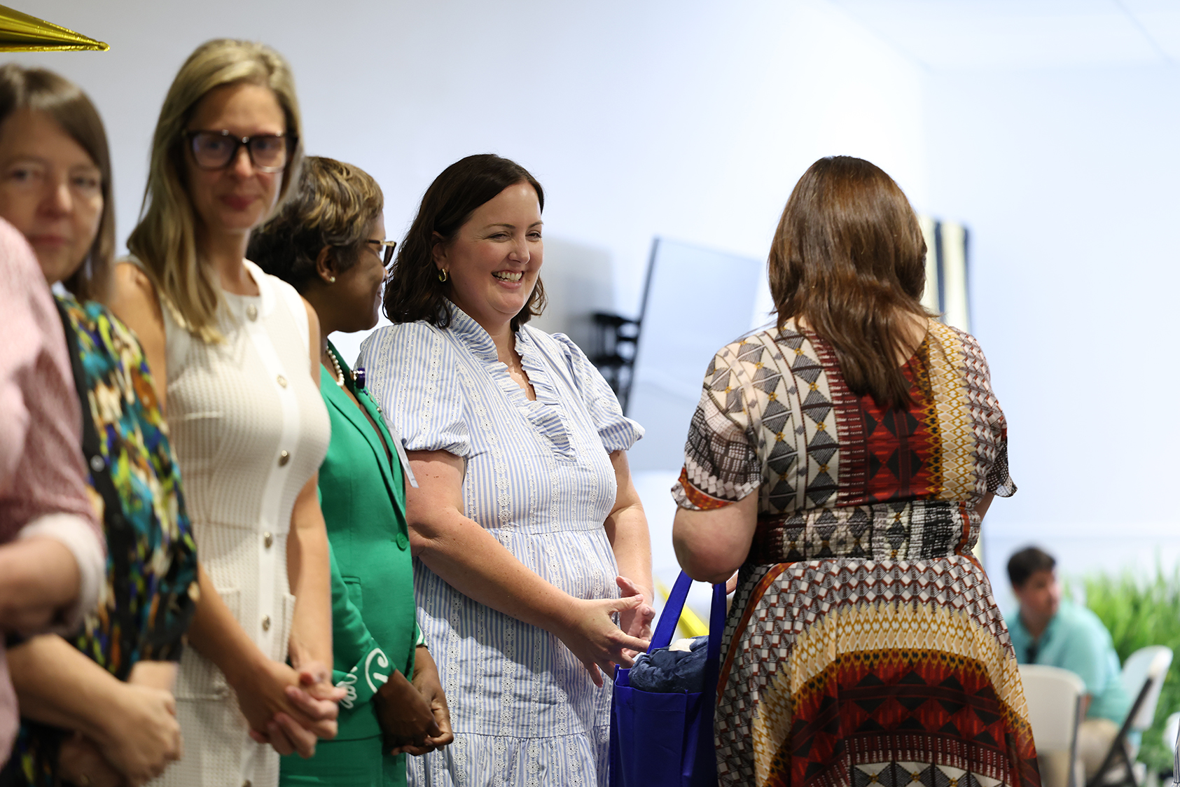 Several women stand together indoors. Most wear dresses, with one carrying a blue bag. Light reflects off the white walls.
