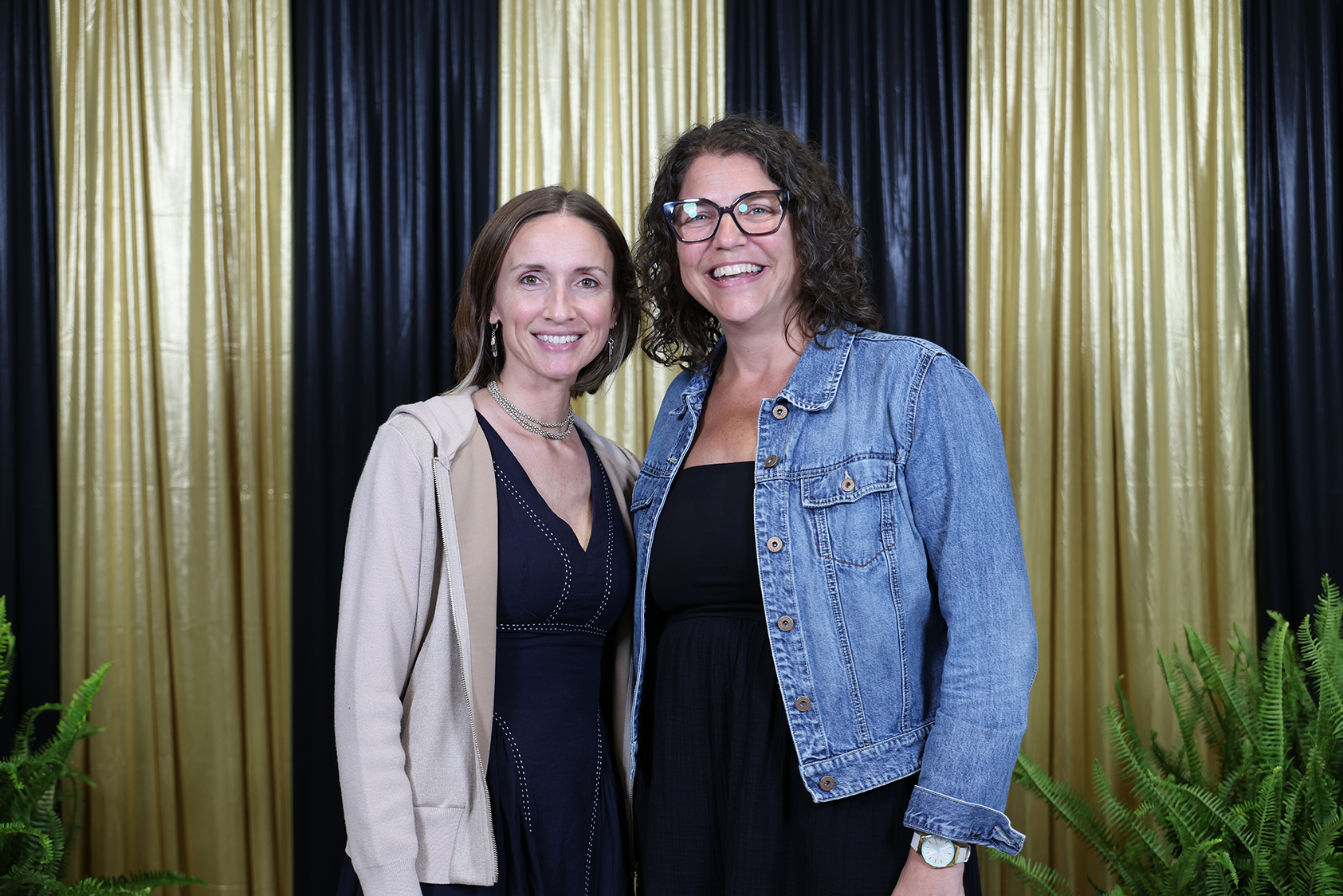 Two women stand against a gold and black backdrop. One wears a denim jacket, the other a dark dress.