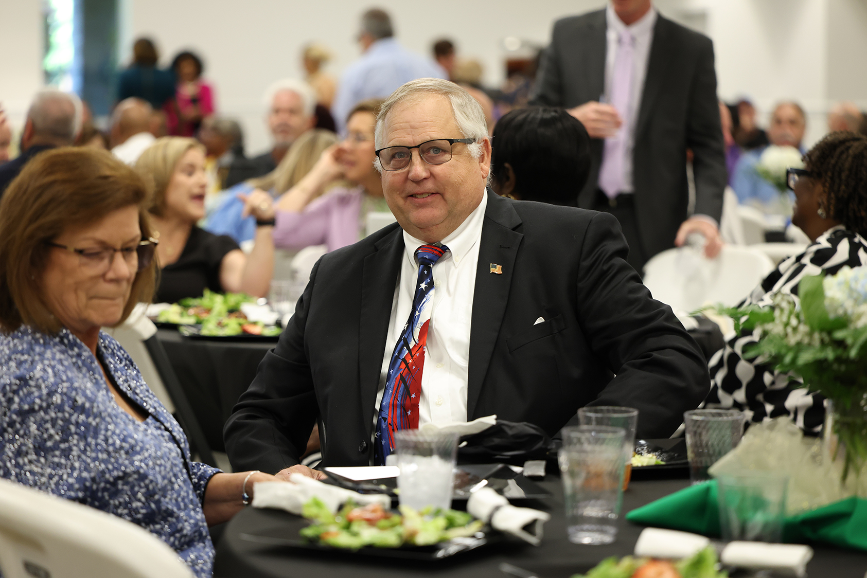 A man in a suit and tie sits at a table with salad, next to a woman in glasses.