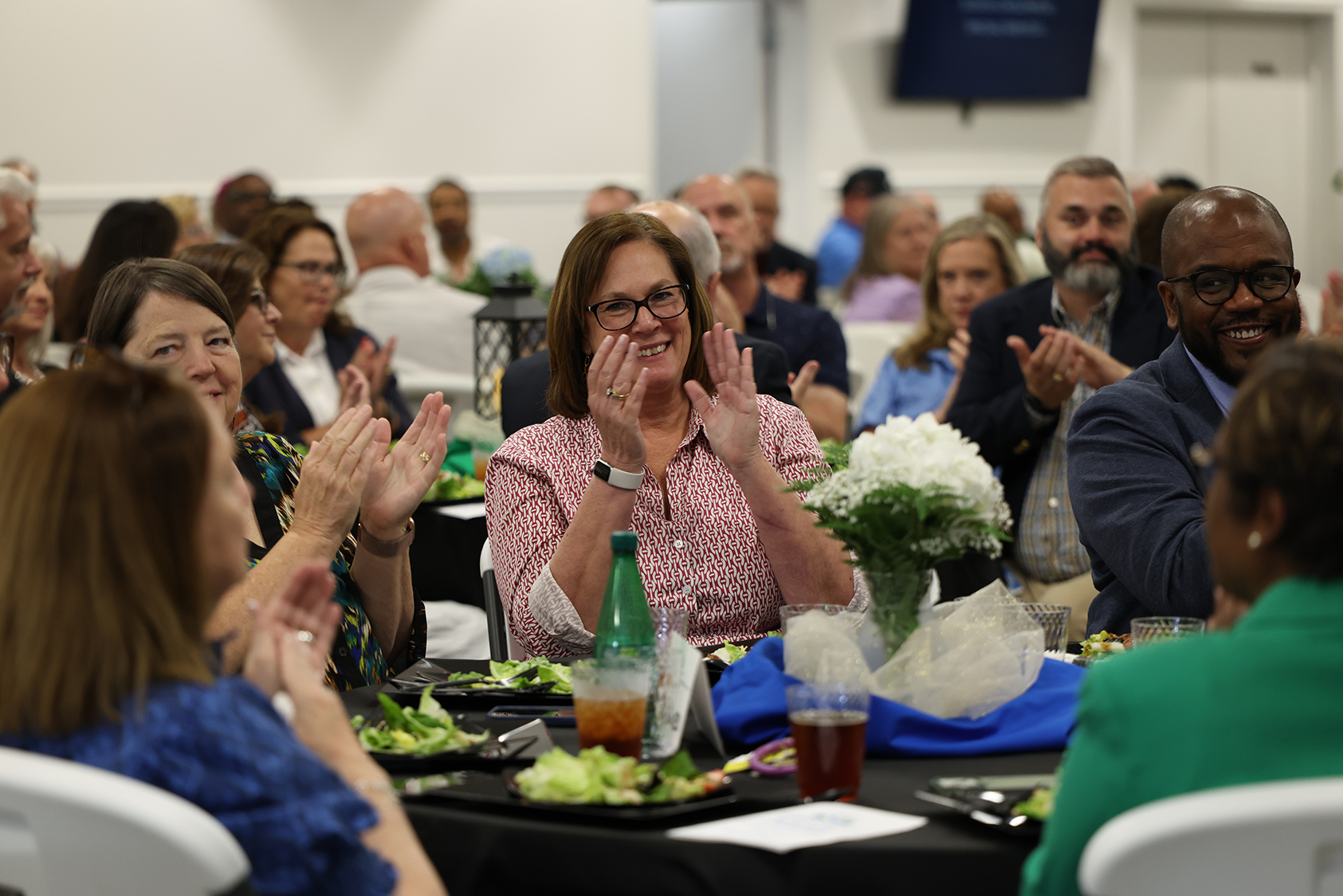 A group of people are clapping in a large room, seated around a long table with plates of food.