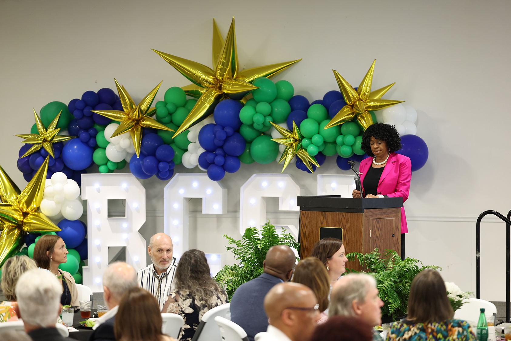A person addresses a seated audience. A podium and balloons in the colors green, white, blue, and gold are behind them.