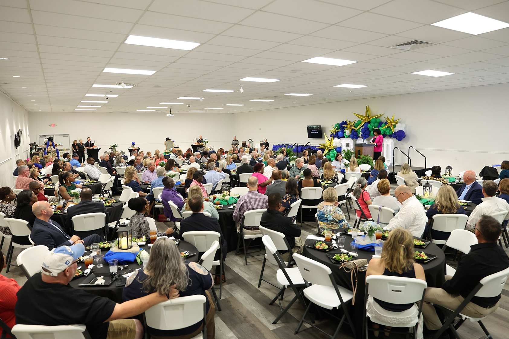 A dining hall with multiple people seated at tables. A stage with a banner and balloons is visible.