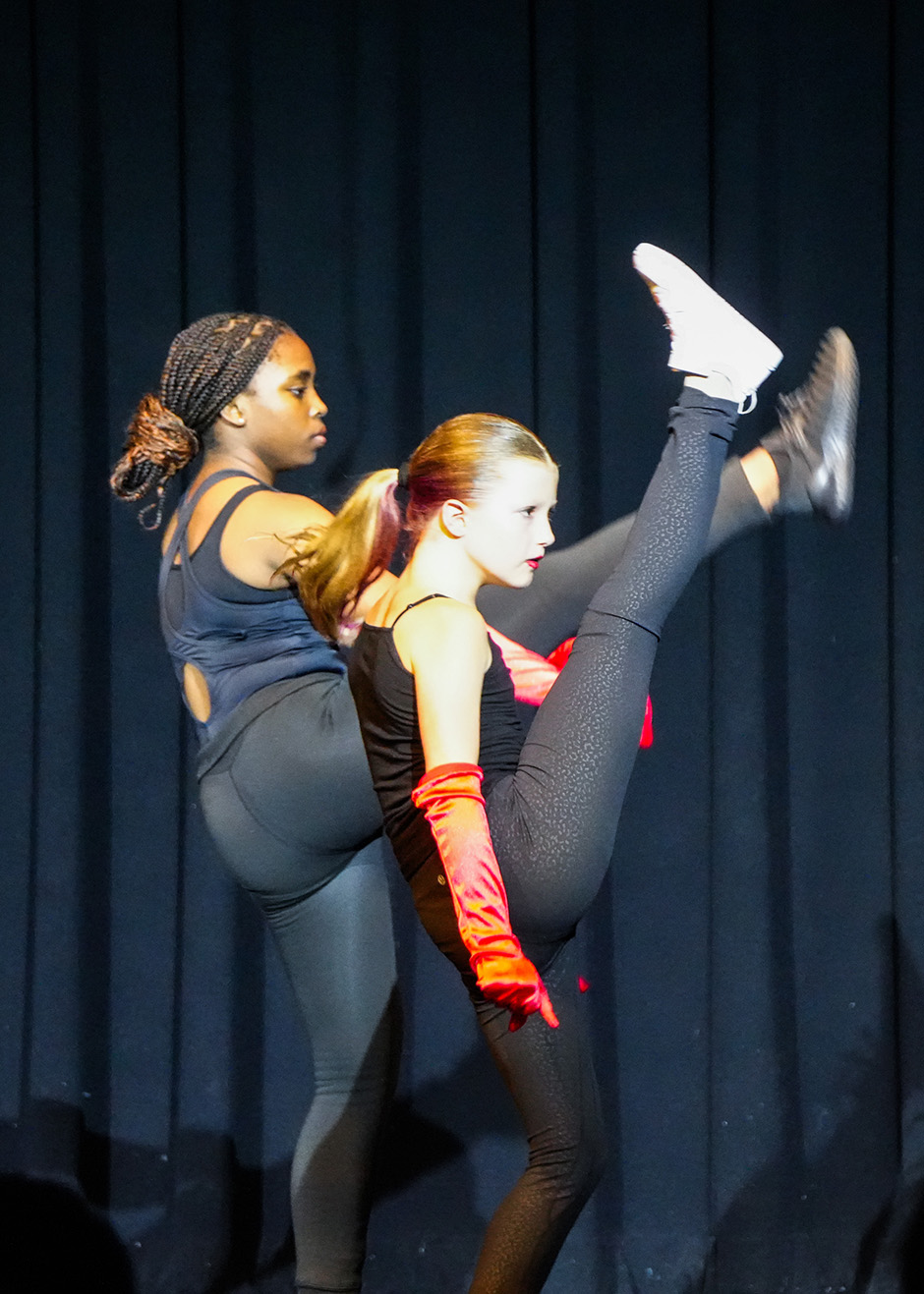 Two women perform a synchronized dance routine on stage, one with legs spread, the other standing with arms behind her back.