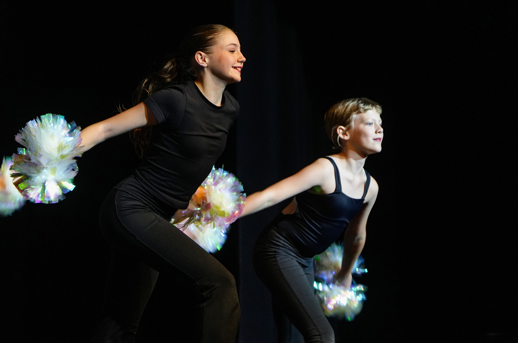 Two young women in black outfits holding pom-poms, performing on stage. One holds pom-poms in both hands, the other in one.