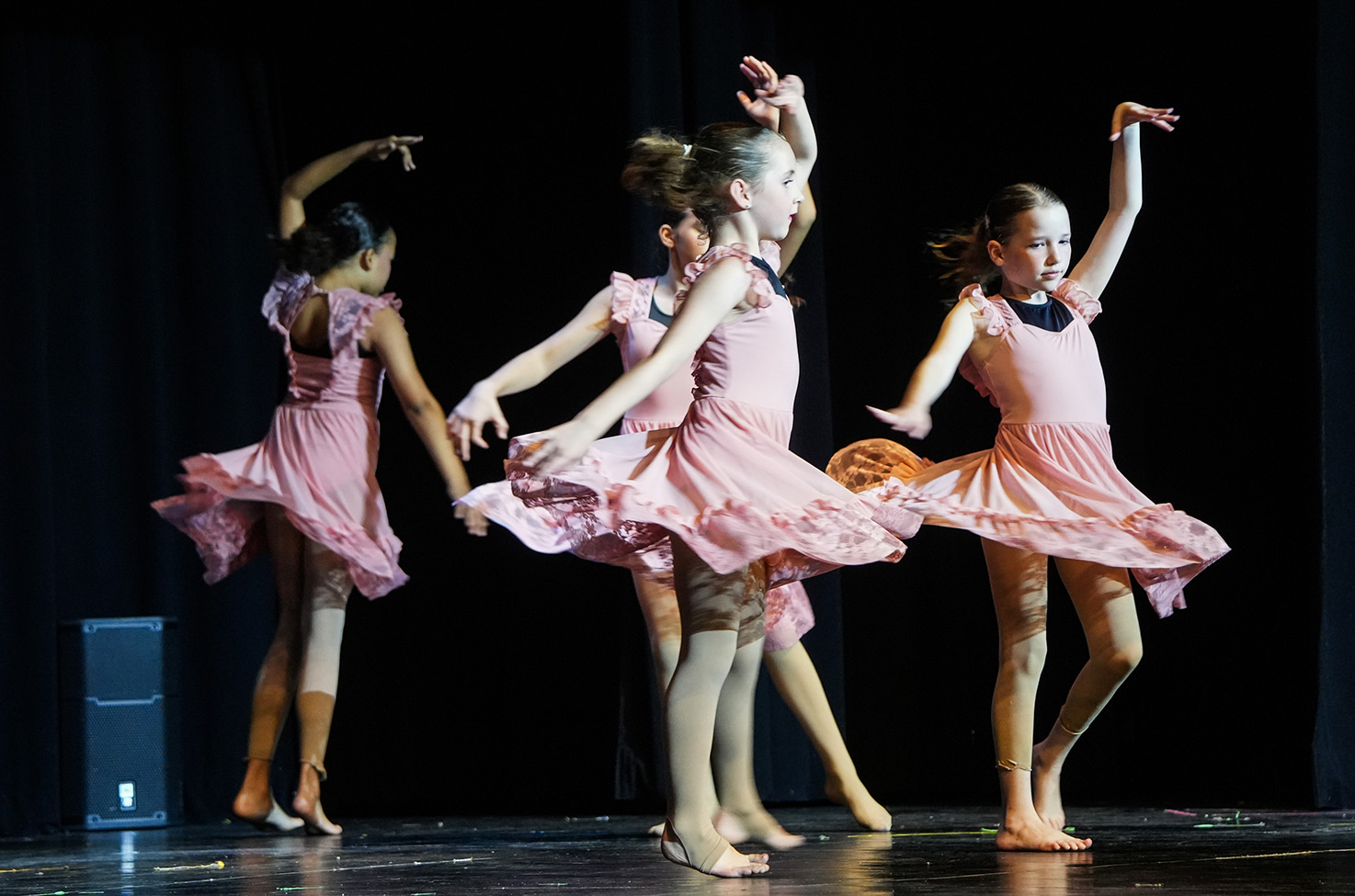 Four young girls in pink dresses perform dance moves on a stage with a black curtain background.