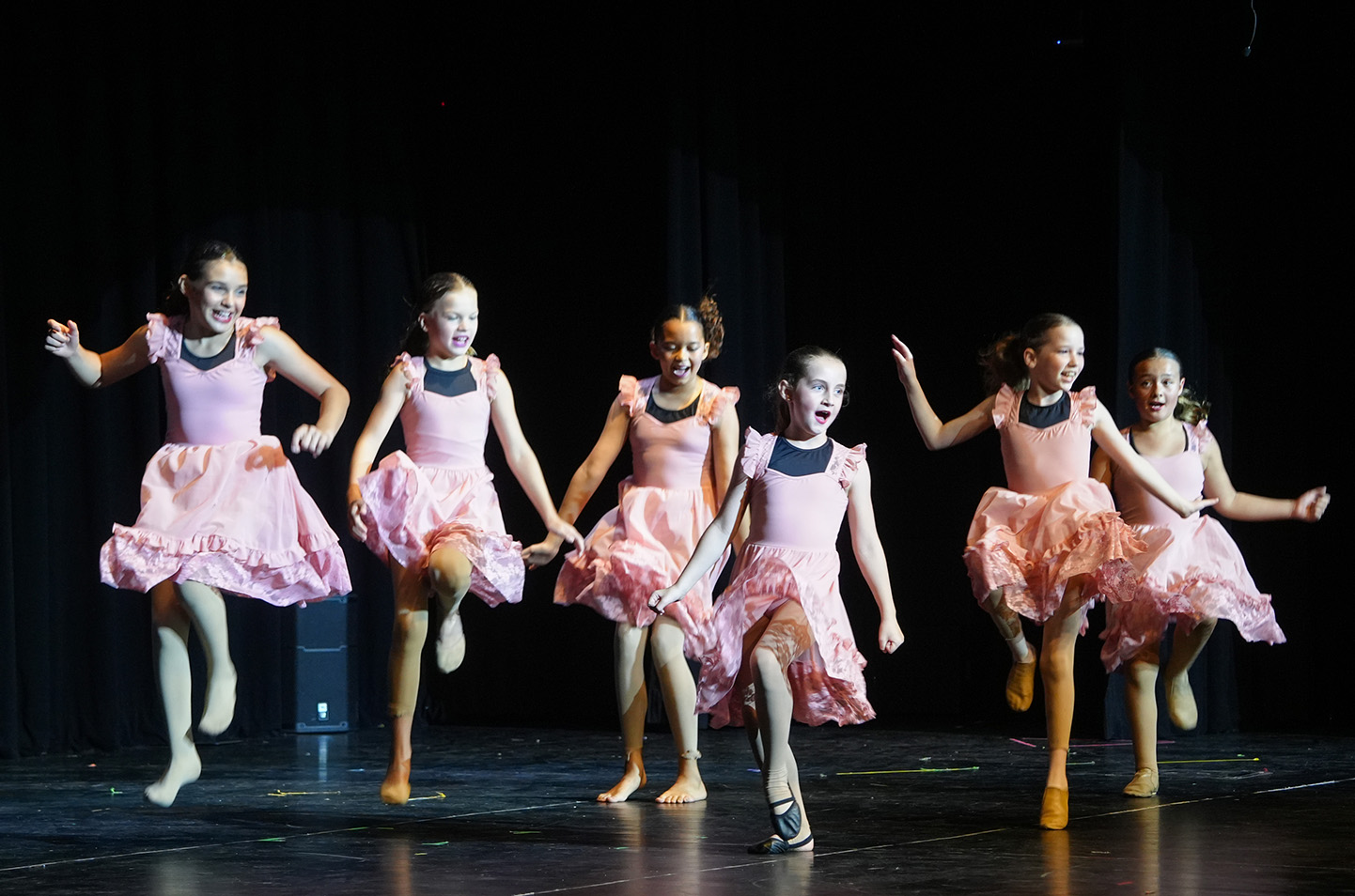Five girls perform a dance routine on a stage, each dressed in pink dresses with ruffles, holding hands.