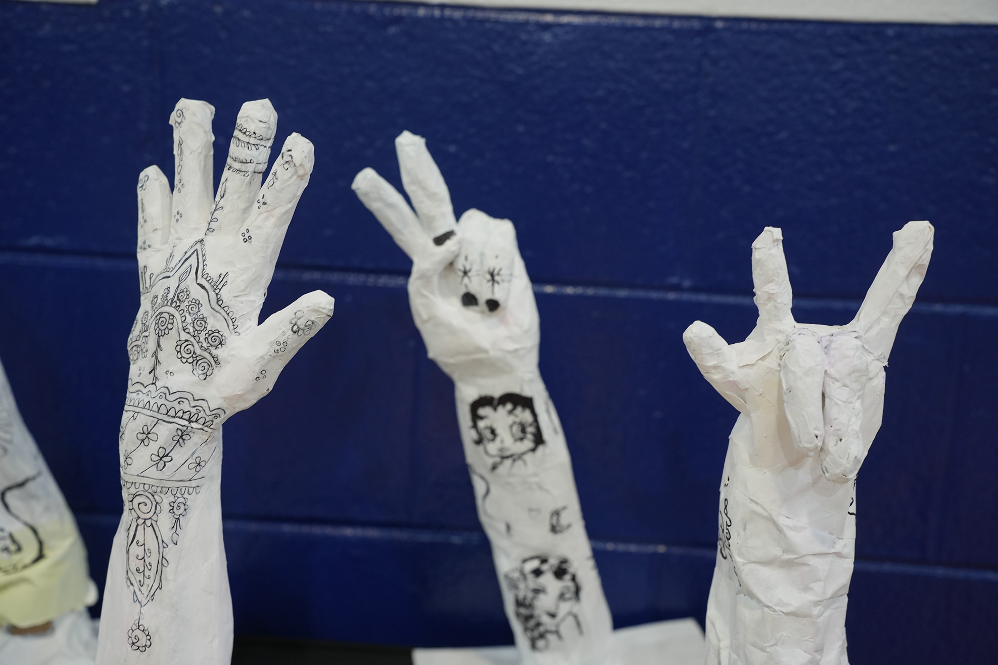 Three white, gloved hands against a blue wall. The left hand has intricate designs, the middle has a drawing, and the right forms a rock sign.