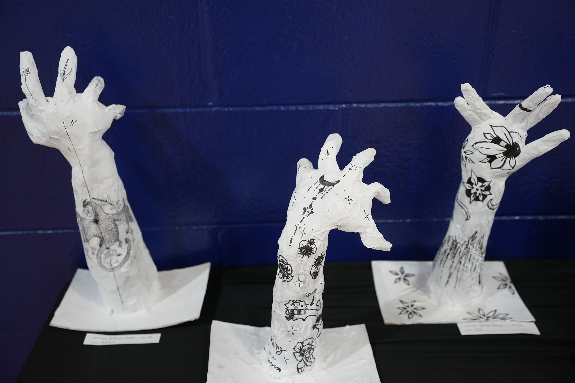 Three hand sculptures are displayed on a black table. Each hand is made of white material, and each is decorated with black designs.