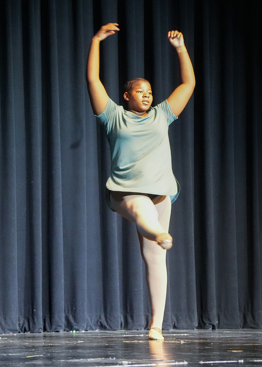 Woman on stage, in the air, wearing green top, pink tights, and ballet shoes, arms and legs spread.