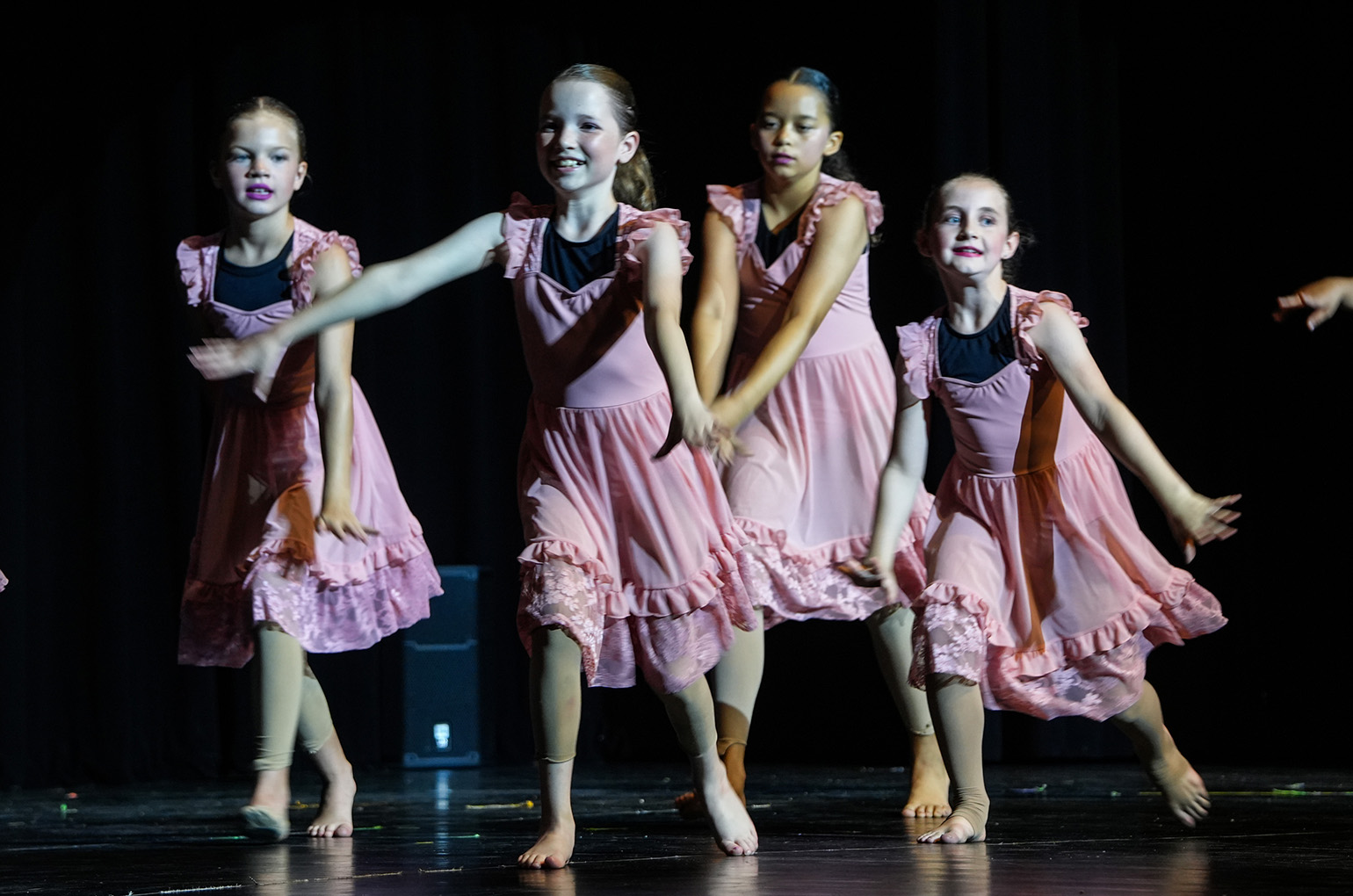 Four young girls in pink dresses perform on a stage. Their hands and feet are in motion.