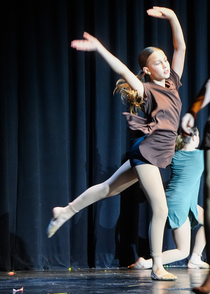 A young dancer wearing a brown top performs a ballet move on a stage.
