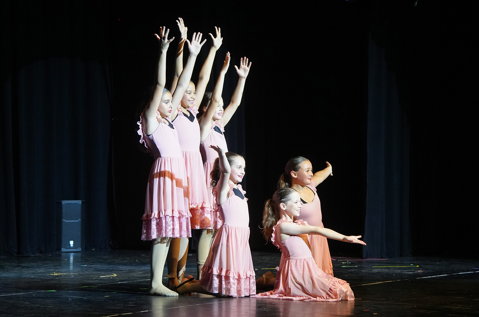 A group of girls in pink dresses on stage, arms raised. The stage has a black backdrop.