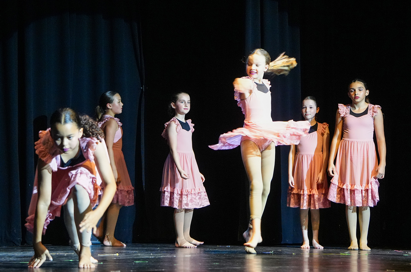On a stage, several young girls in pink dresses perform a dance routine, with one girl leaping and others crouching.