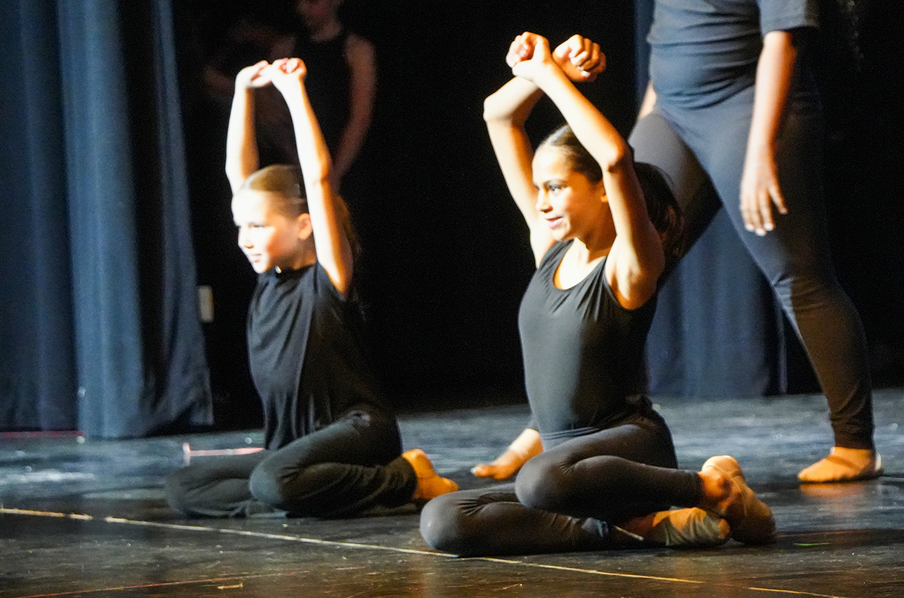 Two dancers in black tops and leggings sit on stage with raised arms, spotlights illuminating them.